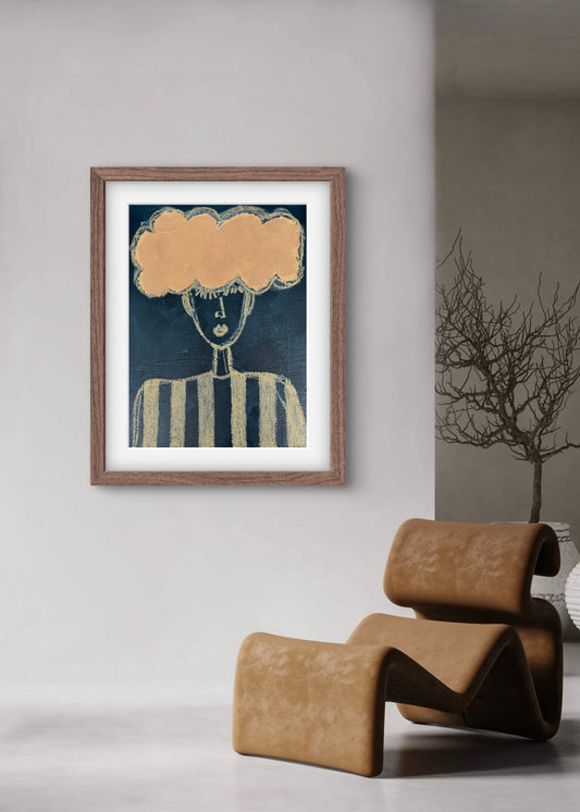 Framed artwork with a cloud above a face on a wall next to a modern brown chair.
